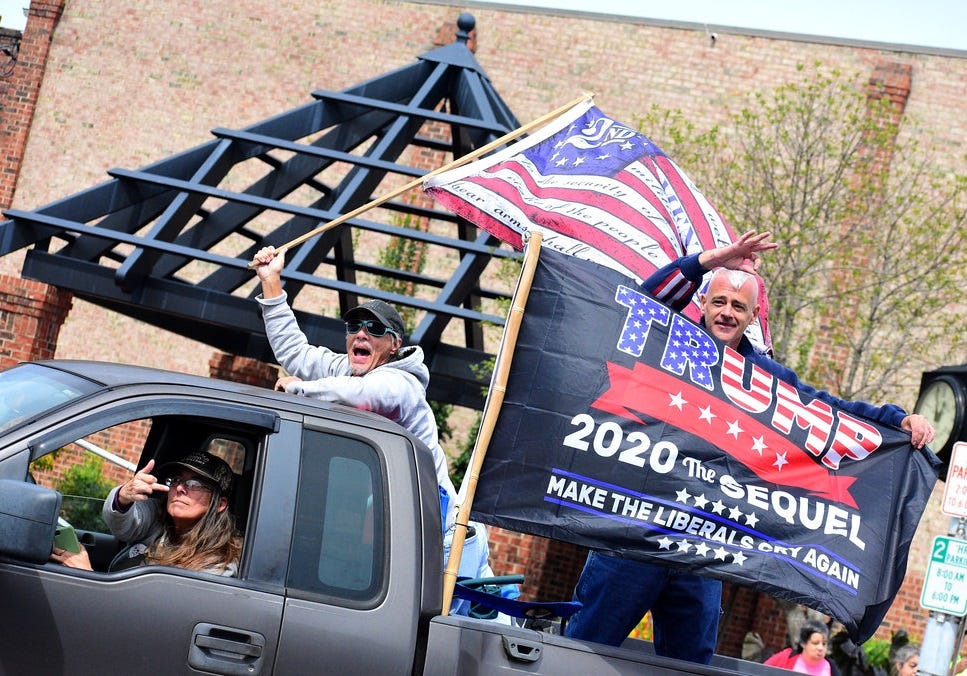 Photo of a grey pickup truck whose driver is flipping the middle finger (not clear whether at photographer or others); in the bed, one shouting man waves a '2nd Amendment' flag (a Betsy Ross flag with '2nd' in the circle of stars and the text of the amendment in old-timey script on the white stripes). Another man gives the white-power 'OK' hand sign and holds up a flag reading 'TRUMP: 2020 THE SEQUEL / MAKE THE LIBERALS CRY AGAIN'