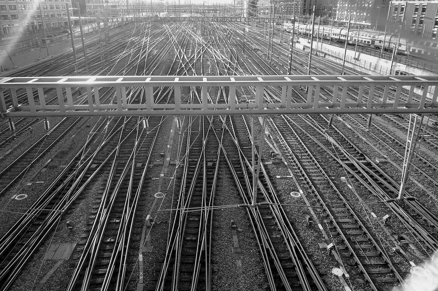 A black and white photo taken from a footbridge overlooking multiple interleaving electrified train tracks.
