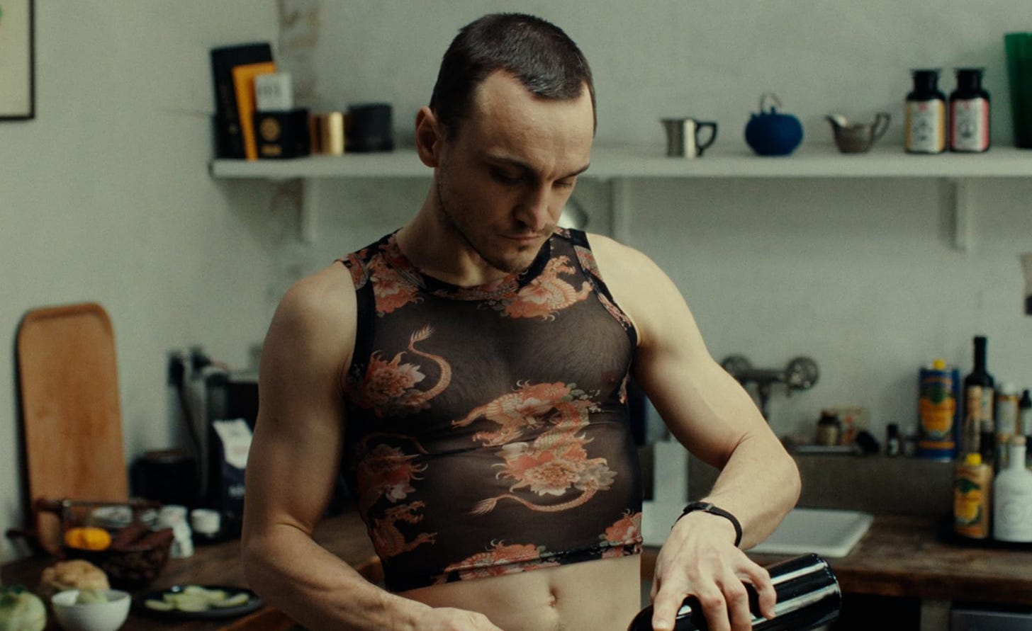 Franz Rogowski wears a mesh crop top with red dragons as he pours wine in "Passages" (2023)