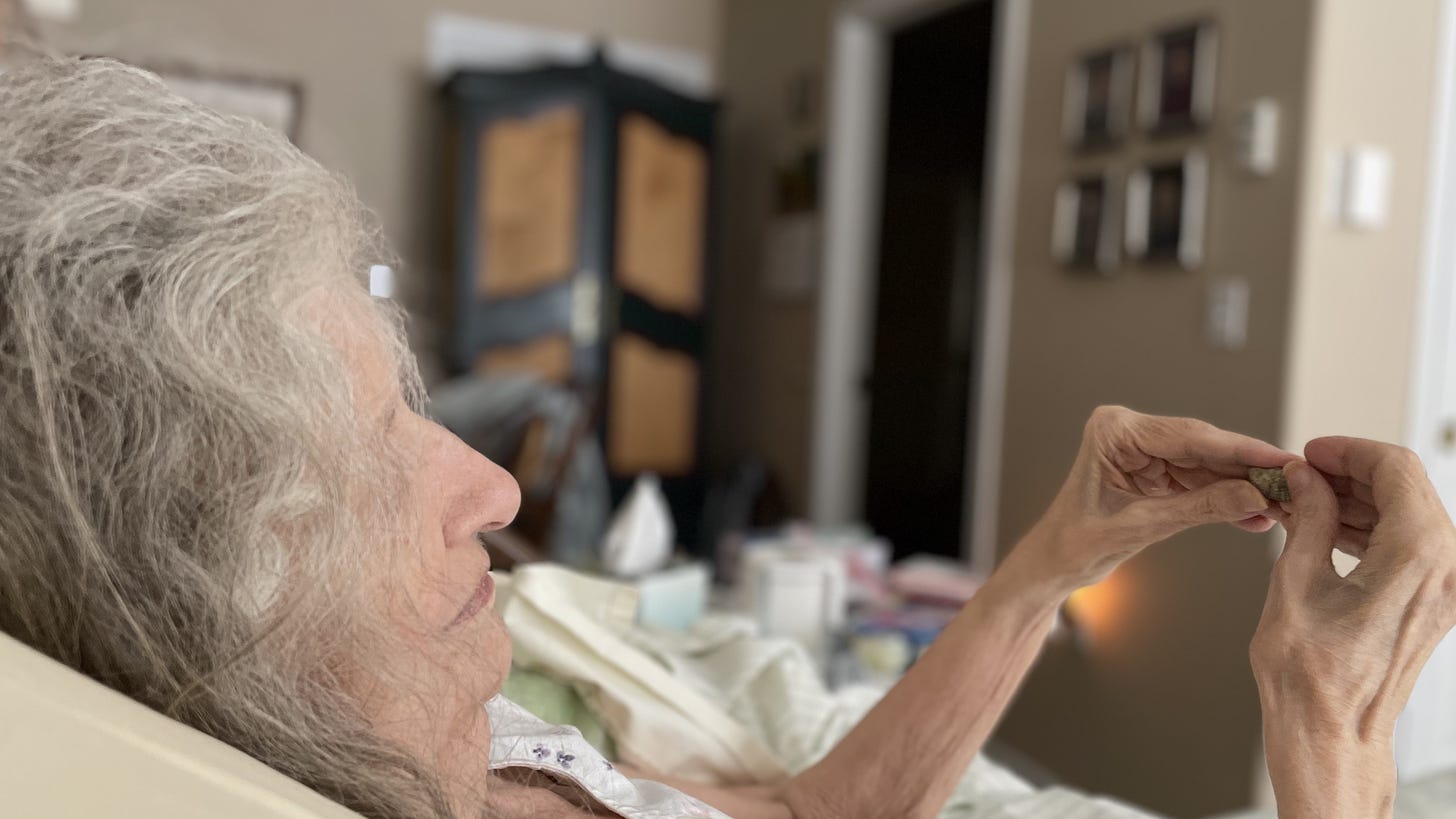 An elderly woman lies in bed and looks at a small fossil she's holding in her hands.