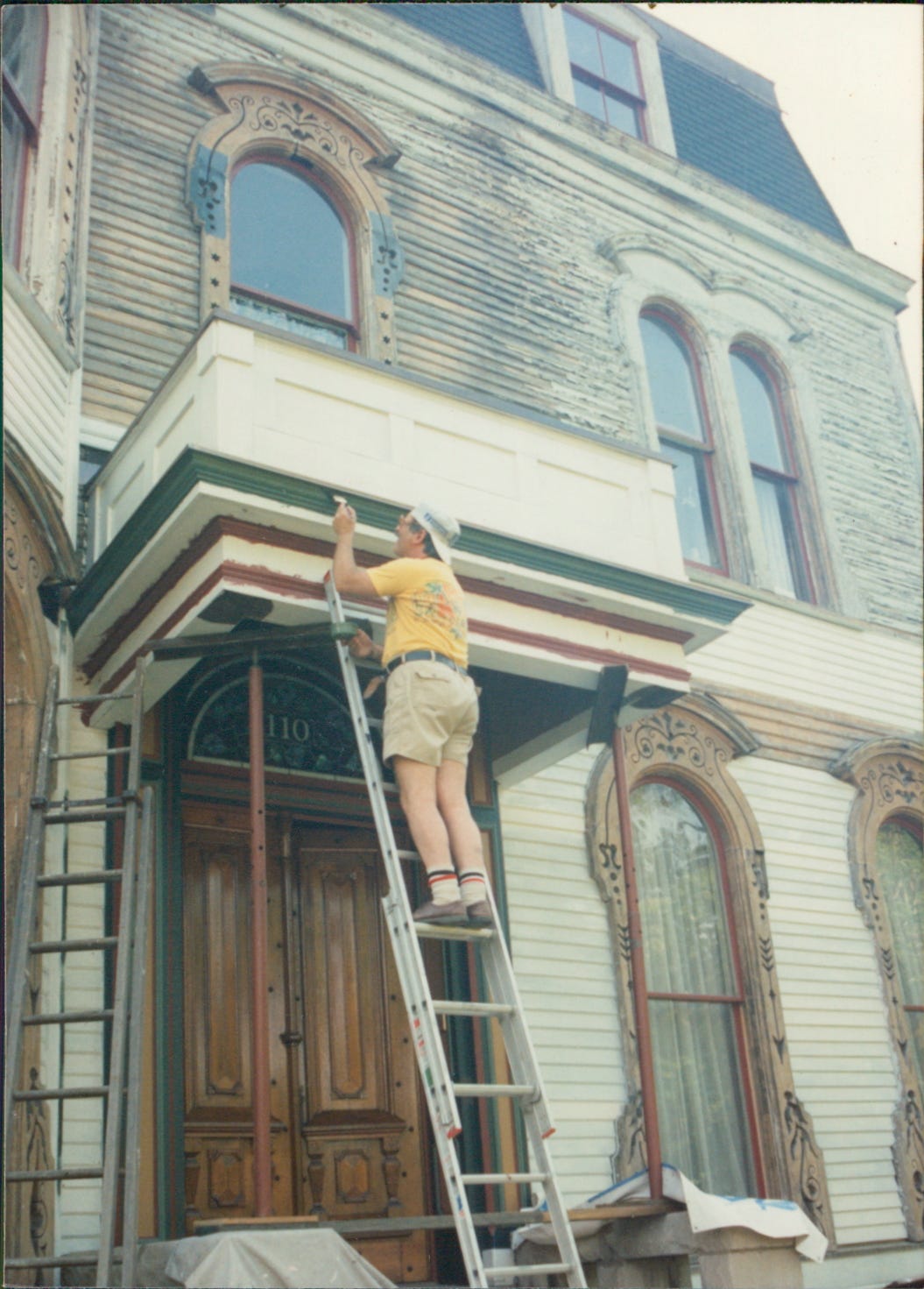 Man on ladder painting the trim on front entrance of old Victorian house. He is wearing a yellow shirt, kahki shorts, and tube socks. The paint is dark green.