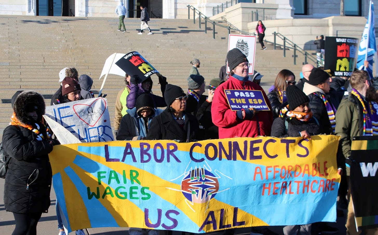 people standing with colorful signs and wearing winter attire hold a banner depicting a blue sky and yellow sun background behind text that reads "labor connects us all" "Fair wages" and. "affordable healthcare" with 4 hands reaching fingertips