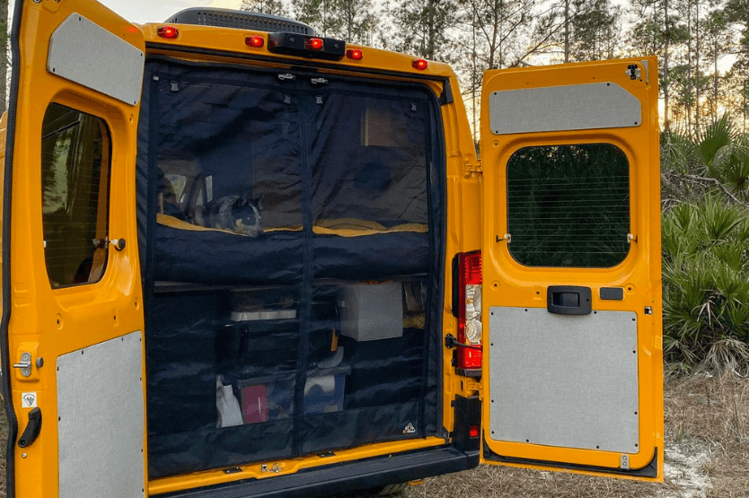 Scout the blue heeler rests on the bed in the back of a converted yellow Ram Promaster van. The van's back doors are open, showing a garage area underneath the bed.