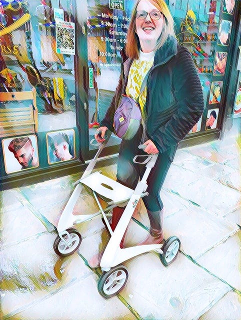 Jane out and about with her rollator. She's smiling, wearing a short brown fluffy jacket, brown boots, yellow top and a purple cross body bag.