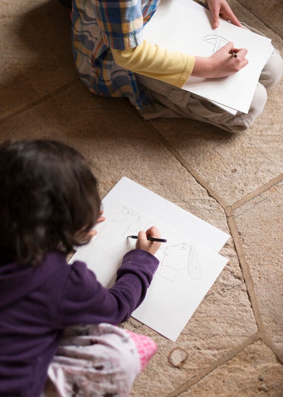 Two children seated on a stone floor and seen from a high angle. One of the figures is cropped by the lower-left corner of the image, and the other by the upper-right corner. Each child draws a picture.