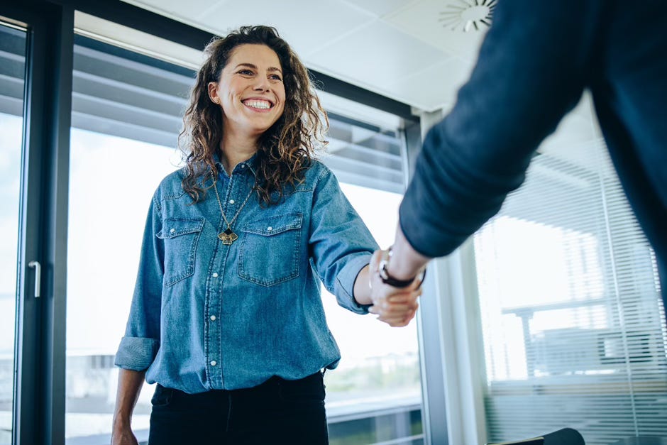 The image shows a smiling woman with curly brown hair wearing a denim shirt. She is shaking hands with a person whose body is partially visible in the frame. The woman appears to be in a professional or business setting, such as an office or workplace, with windows visible in the background. Her warm, friendly expression and the handshake gesture suggest a positive interaction or greeting between the two individuals.