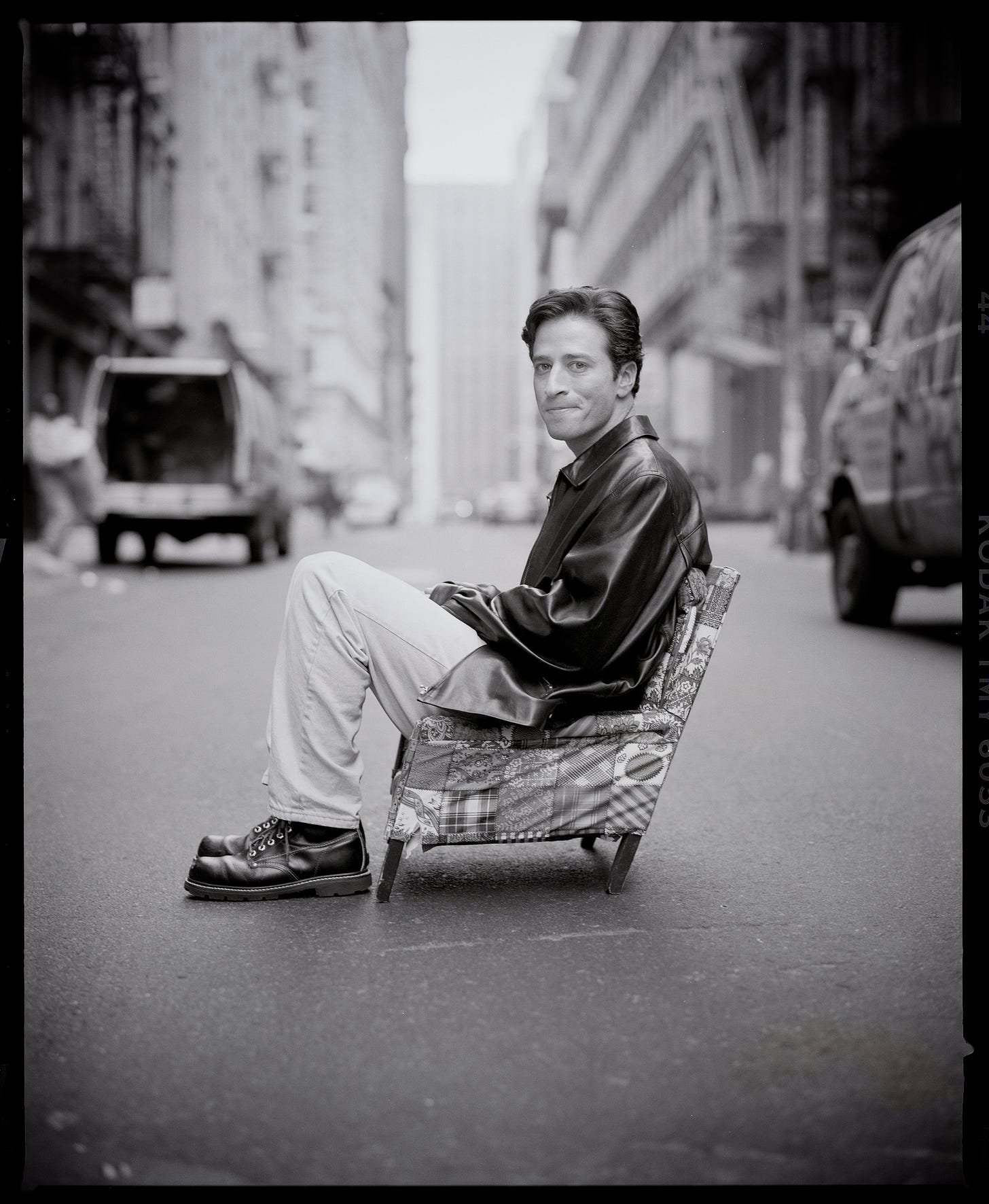 Jon Stewart sitting in a tiny chair in the middle of the street.