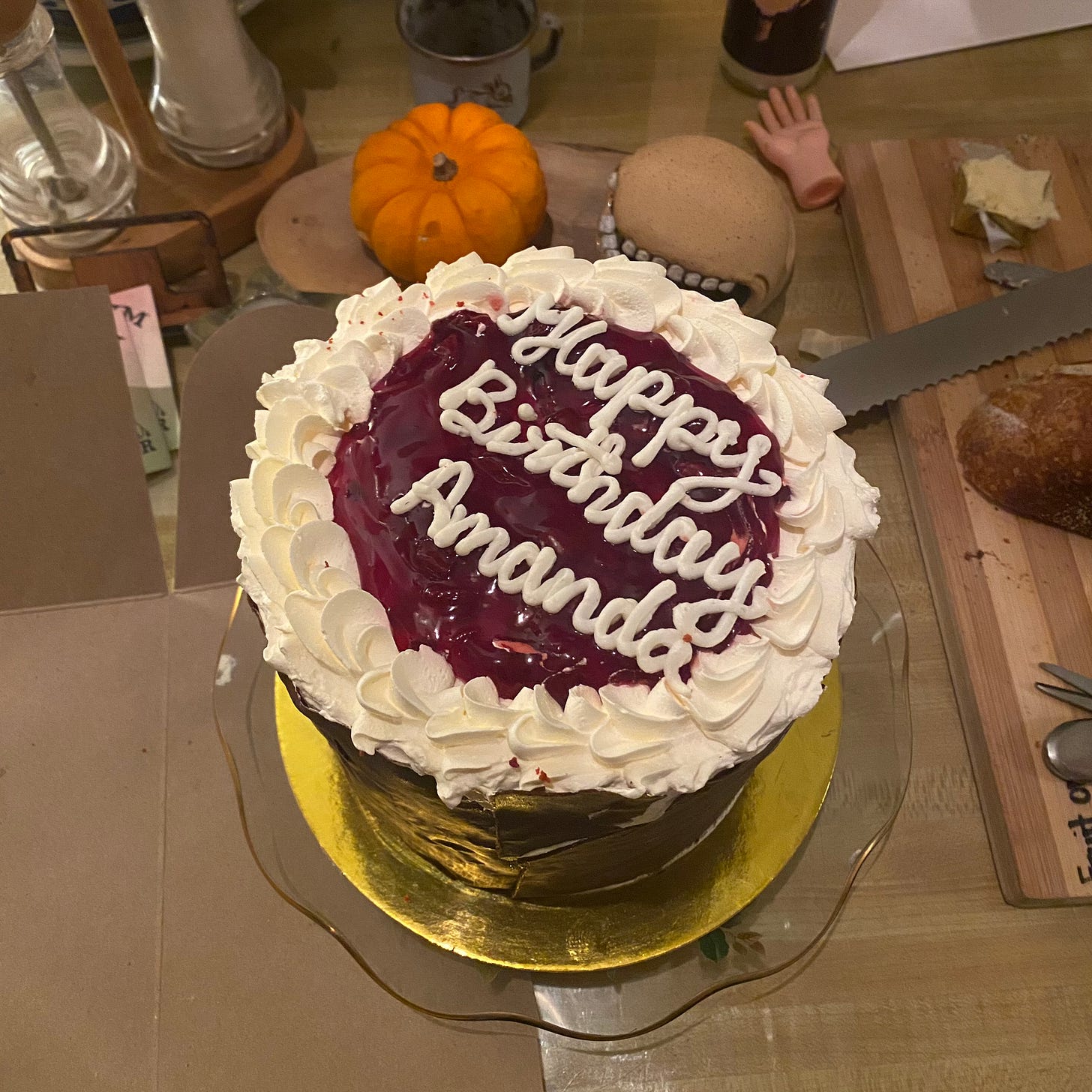 A tall 6" black forest cake with a chocolate bark shell around the outside, the top covered in piped whipped cream and sour cherry preserves. Writing in white reads 'happy birthday Amanda' on top of the cake. There are some items on the table around the cake: a mini pumpkin, a breadboard with a knife and a loaf of sourdough on it, a doll's hand, a sculpture with teeth, a candle, a salt & pepper duo.