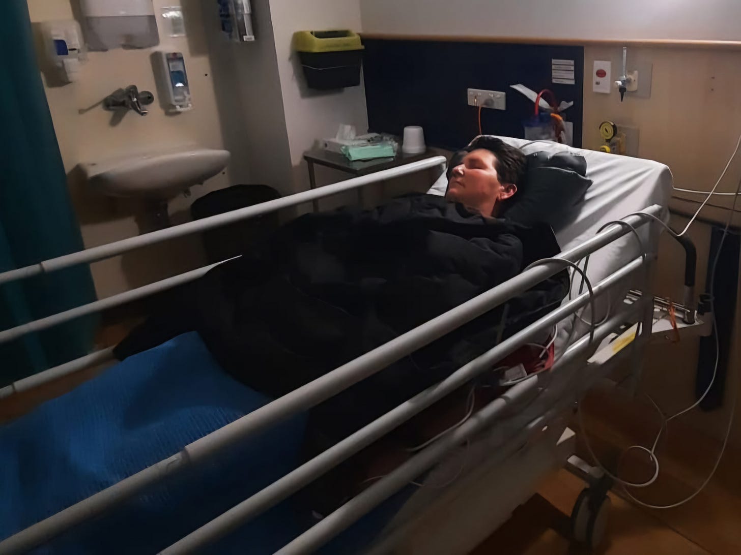 Kathryn is resting in a hospital bed in an Emergency Deparment, covered by a black jacket and blue blanket. There's a sink n the backround and the usual hospital cords and equipment.
