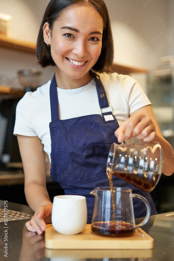 Vertical shot of smiling cute barista girl, pouring filter coffee, making order for cafe client, wearing blue apron