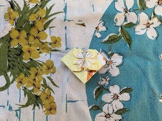 A yellow origami heart sits on a turquoise and white tablecloth of yellow and white flowers.