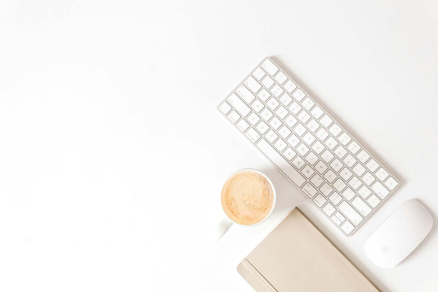 white desk top with wireless keyboard and wireless mouse near cup of coffee and beige notebook