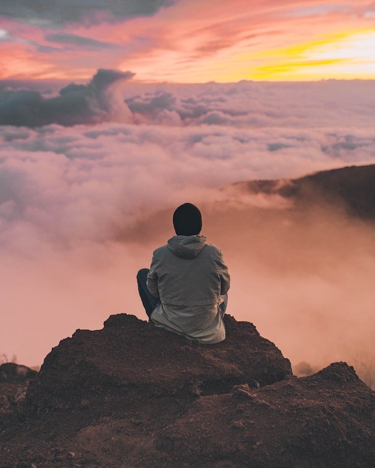 A person medidates on a cliff watching the sun rise over the clouds