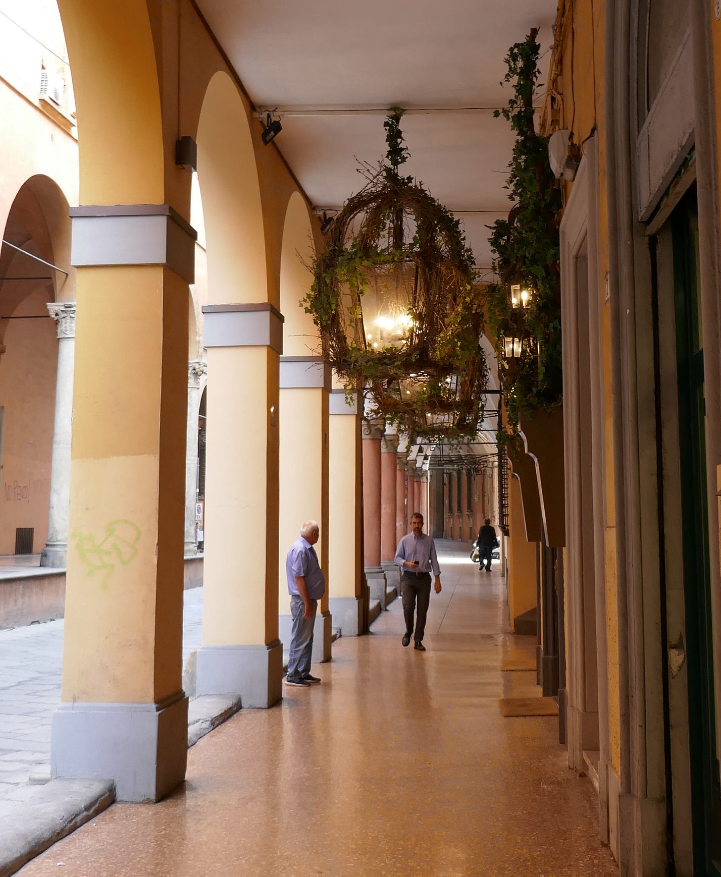 A few pedestrians walk through a portico with yellow columns and elaborate chandeliers with creeping vines.