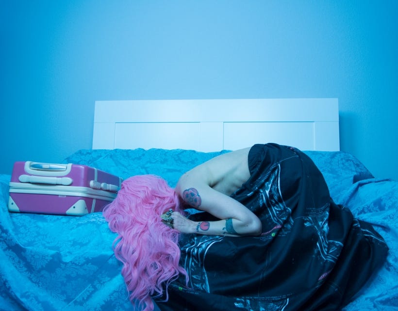 Karrie in the fetal position, face down on a blue taffeta couch, with long, bubble-gum pink hair flowing over the cushions. She is wearing a long, flowing skirt printed with her c-spine MRI. Next to her head on the couch is a pink suitcase.