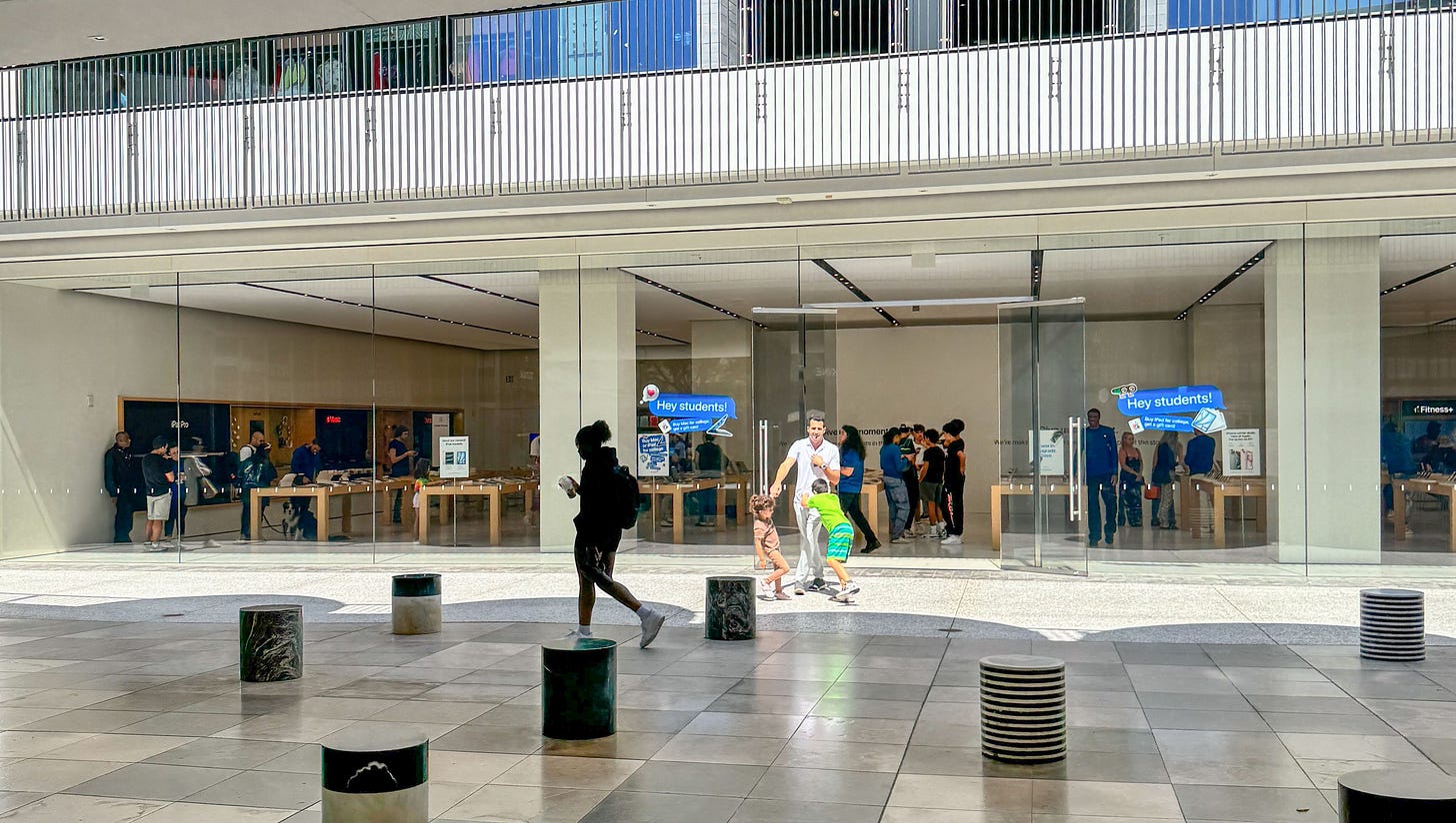 A large white barricade surrounds the Forum at Apple Century City. People are walking in front of the store.