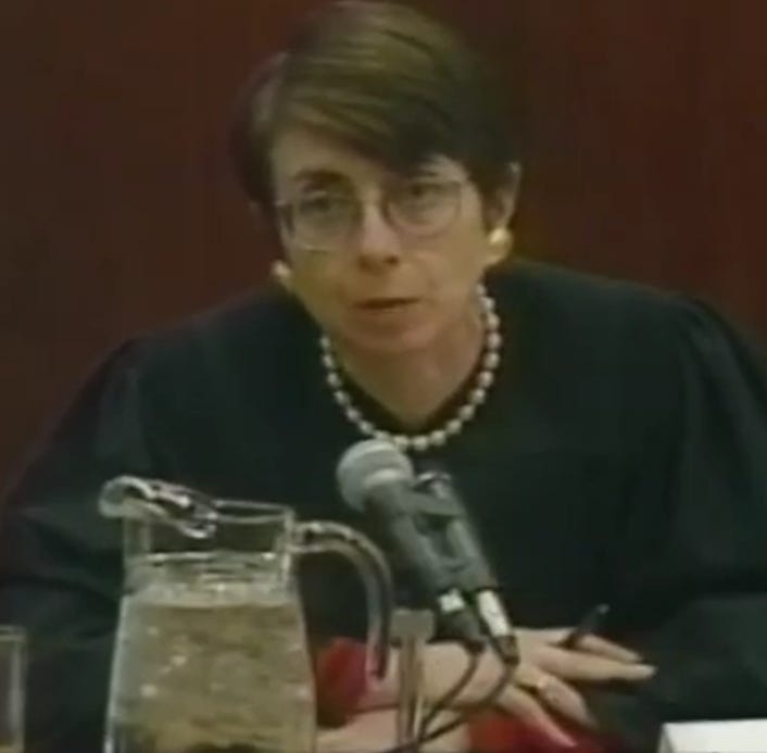A seated woman in a black robe with short brown hair and glasses, behind two microphones and a pitcher of ice water
