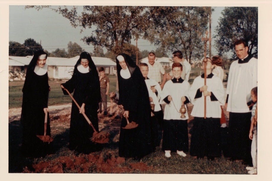 Three nuns in black habits, three altar boys, and priest and some lay people gathered around a groundbreaking for a new church