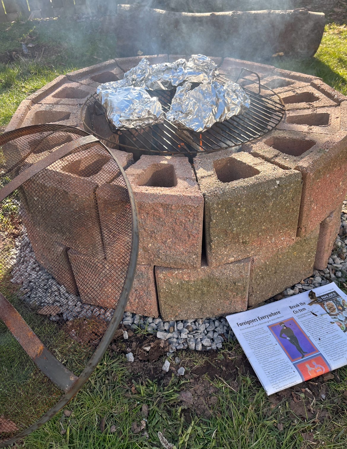 A brick fire pit coveredd in a barbeque grate with packages wrapped in foil roasting on top