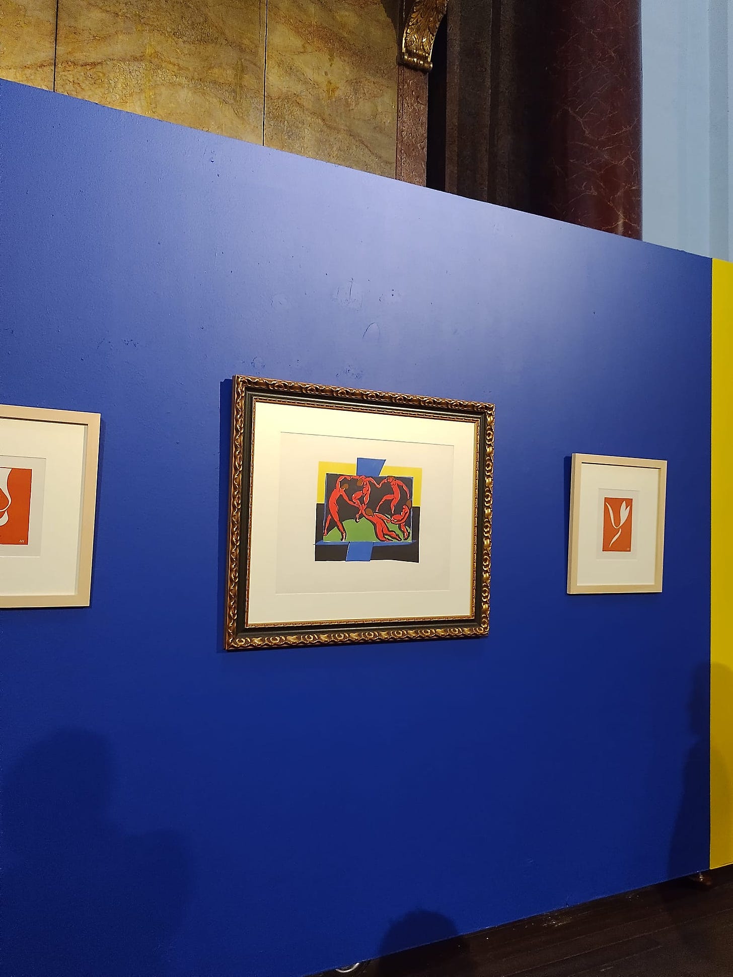 parede azul onde estão pendurados três quadros, os dois das extremidades são colagens representando flores na cor laranja. O do meio é a dança, com figuras humanas nuas dançando em roda.