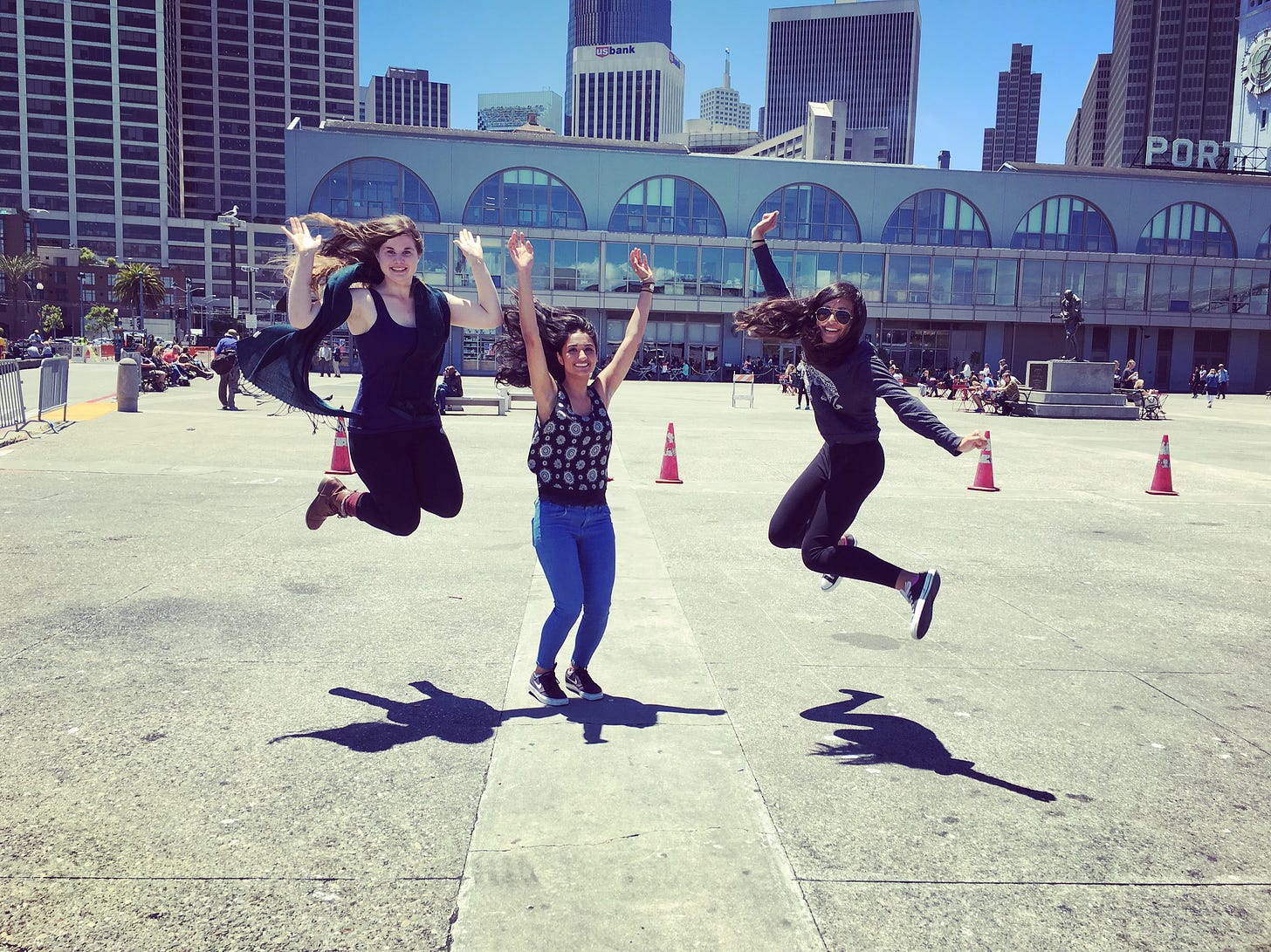 Three women jump in front of San Francisco's Ferry Building. The woman on the left has long brown hair and wears dark clothing. The woman in the middle stands mid-jump with her hands up and wears a dark top with blue jeans. The woman on the right wears sunglasses and dark clothing.