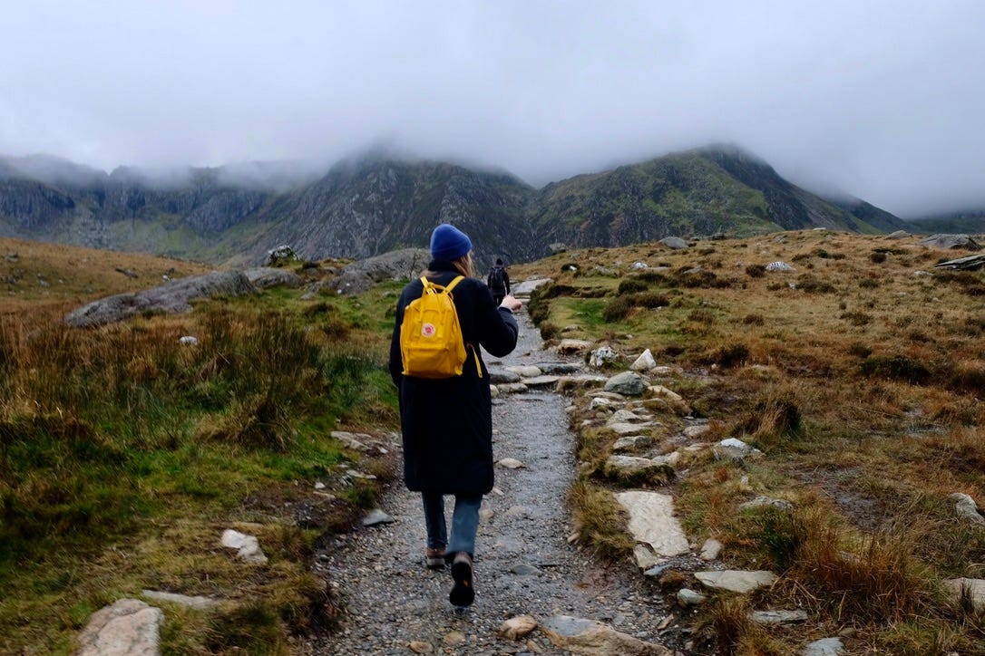 A woman in a blue hat, black coat, and yellow backpack walks on a natural hiking trail towards a rocky mountain shrouded in mist.