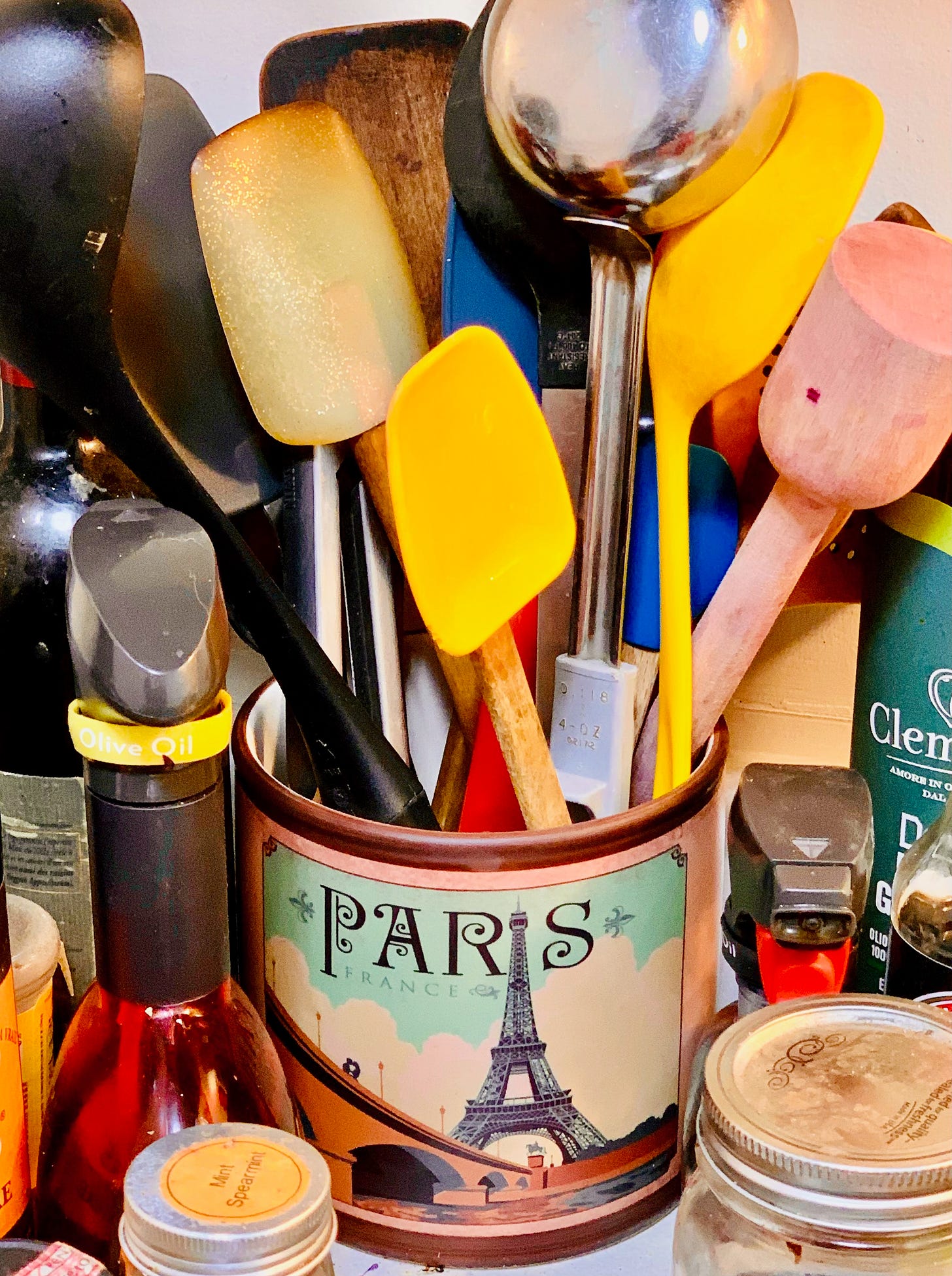 A crock on whose side is an image of a bridge over a river (the Seine, presumably) and in the background the Eiffel Tower, whose tip serves as the "i" in the word "PARIS." The crock is filled with utensils — 3 silicone spatulas (yellow, blue, and cream-colored), a kraut pounder, 3 wood spatulas, several wood spoons, a steel ladle with a grey handle, yellow plastic large spoons including a large round perforated spoon.