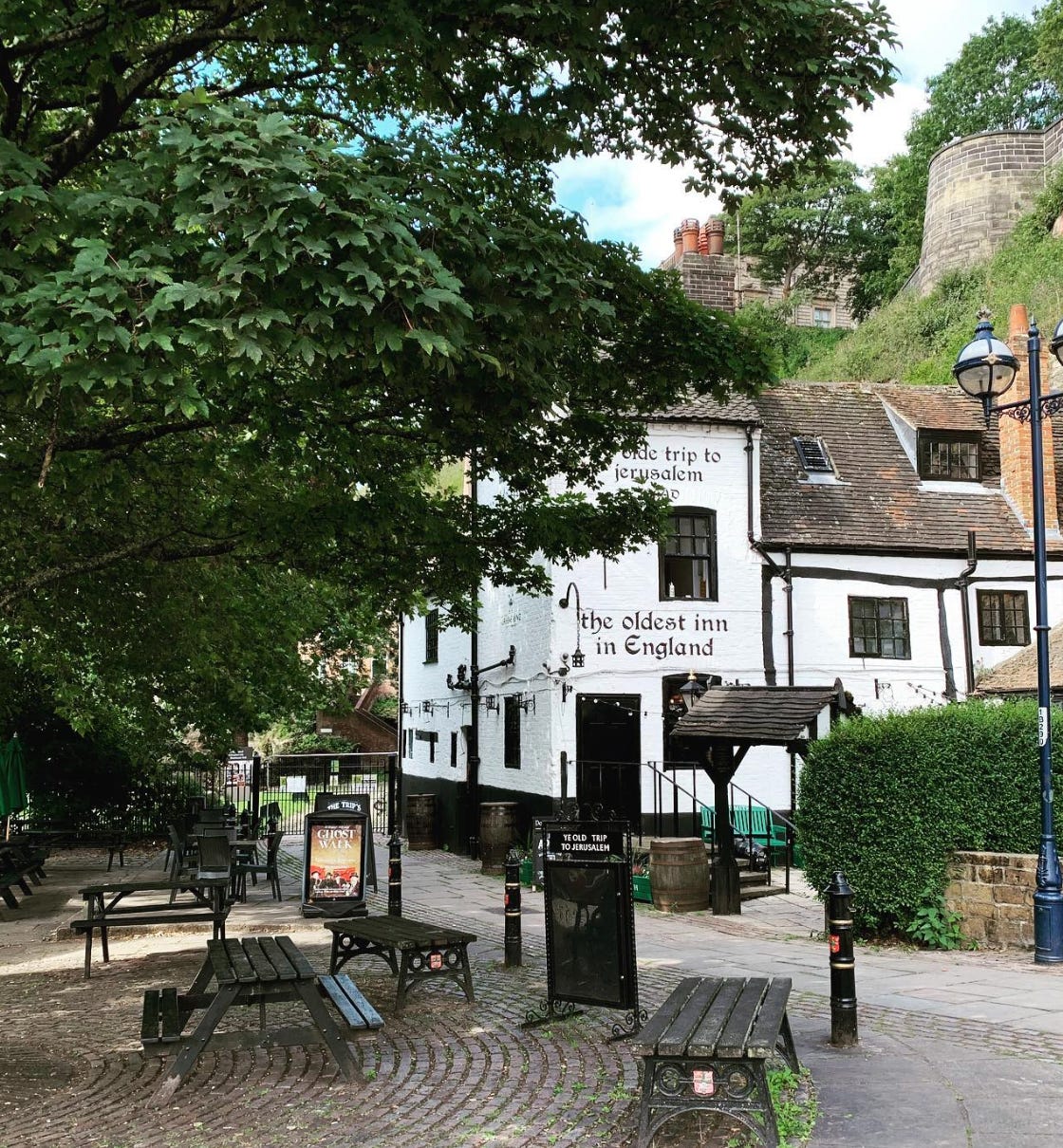 A courtyard with several wooden picnic tables and a big tree leading to a white building with "ye olde trip to Jerusalem" painted on the side