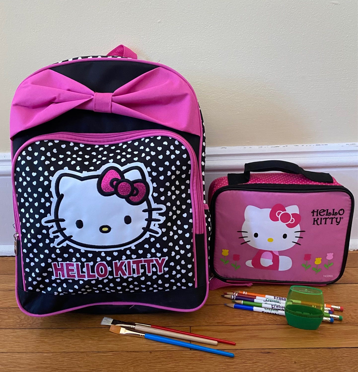 Signature black, pink, and white Hello Kitty backpack and lunch bag, sitting on a hardwood floor, with paintbrushes and colored pencils in foreground.