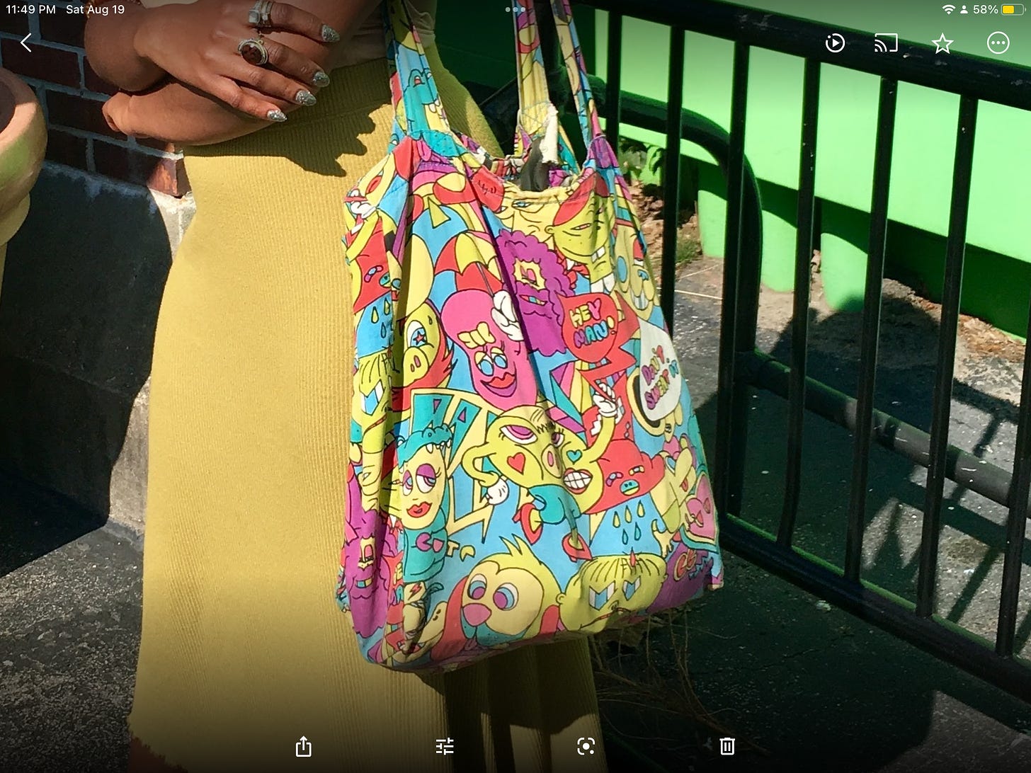 a screenshot of a young Black woman’s arms crossed in front of her waist. she is wearing sparkly nails, rings, and a green skirt with a colorful large tote bag on her arm.