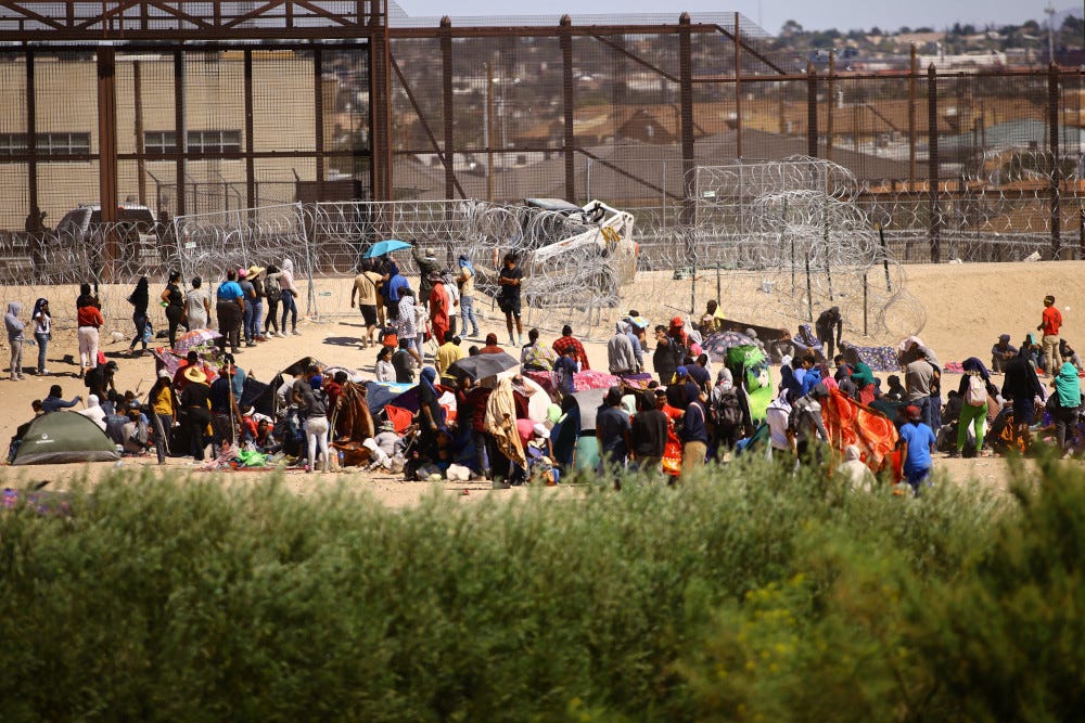 Many people with tents and umbrellas stand on desert land in front of barbed wire structures in front of the rust-colored U.S. border wall