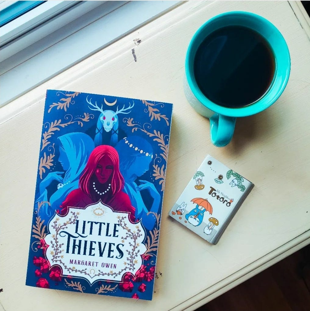 On a light yellow table by a window sits the book Little Thieves by Margaret Owen, a blue mug of coffee, and a pack of Totoro post its.