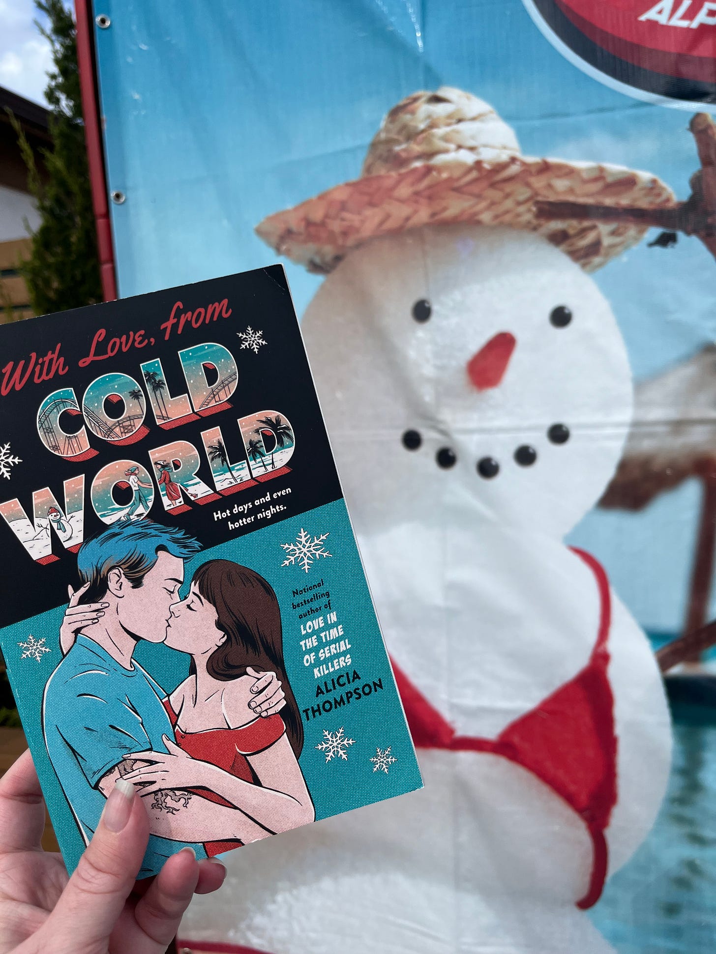 A copy of the teal cover copy of WITH LOVE, FROM COLD WORLD in front of a poster that shows a snowman with a bikini top on