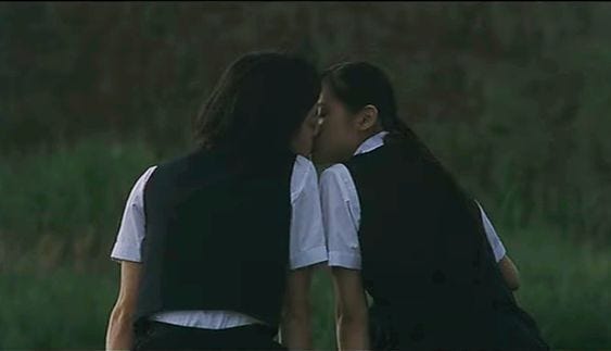 This may contain: two women kissing each other in front of tall grass and trees, with one woman wearing a white shirt and black vest