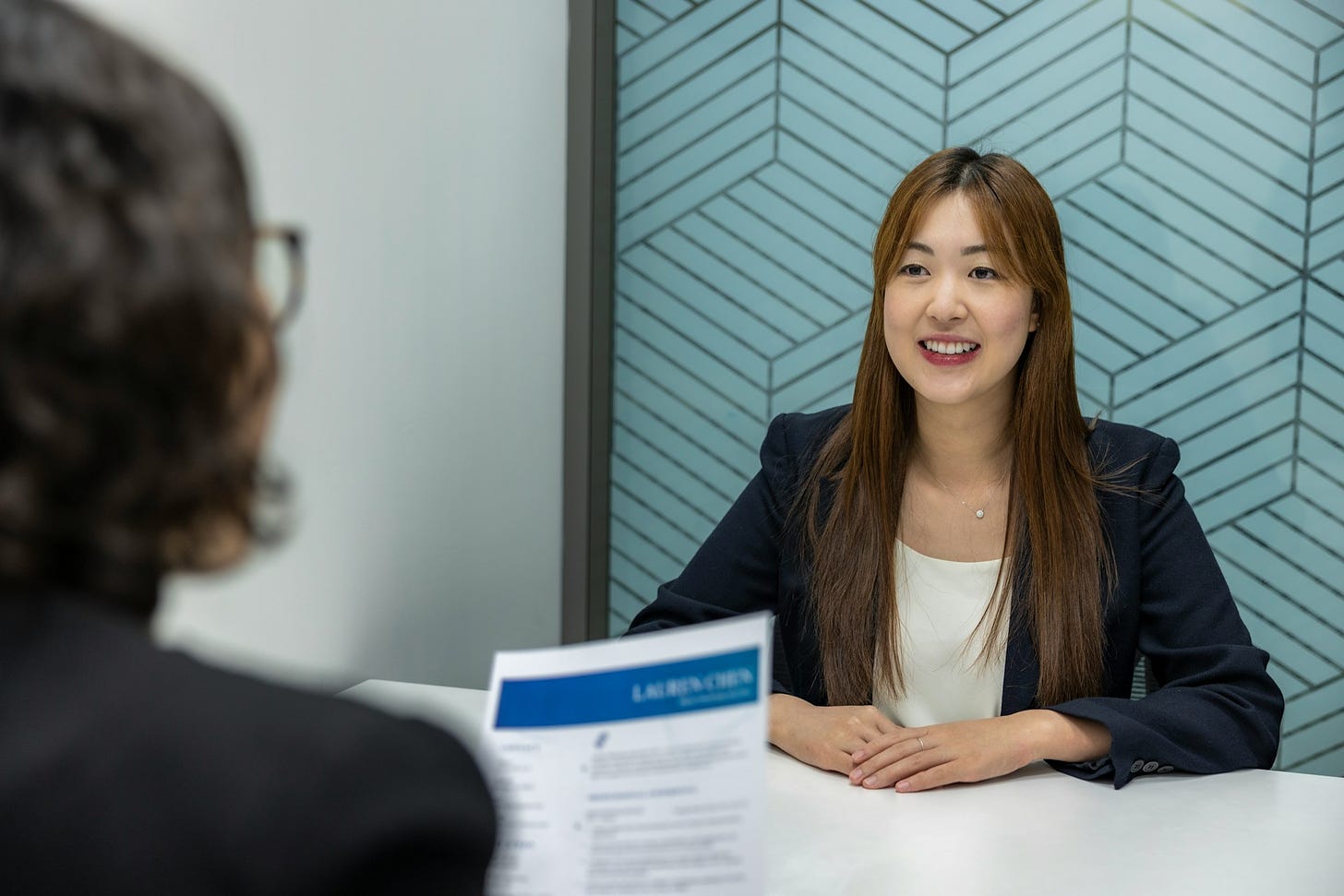 An over-the-shoulder view of an interviewer, holding a resume, with a woman facing her across a table.