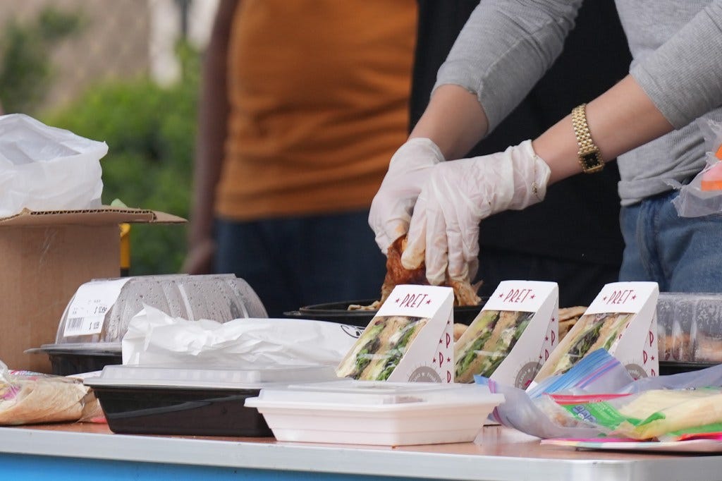 Sandwiches and food containers on an outdoor table with a pair of gloved hands in the background
