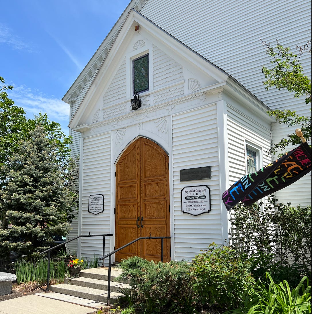 Old clapboard church, white with oak doors, a stained glass panel over the door, and a pride flag. 