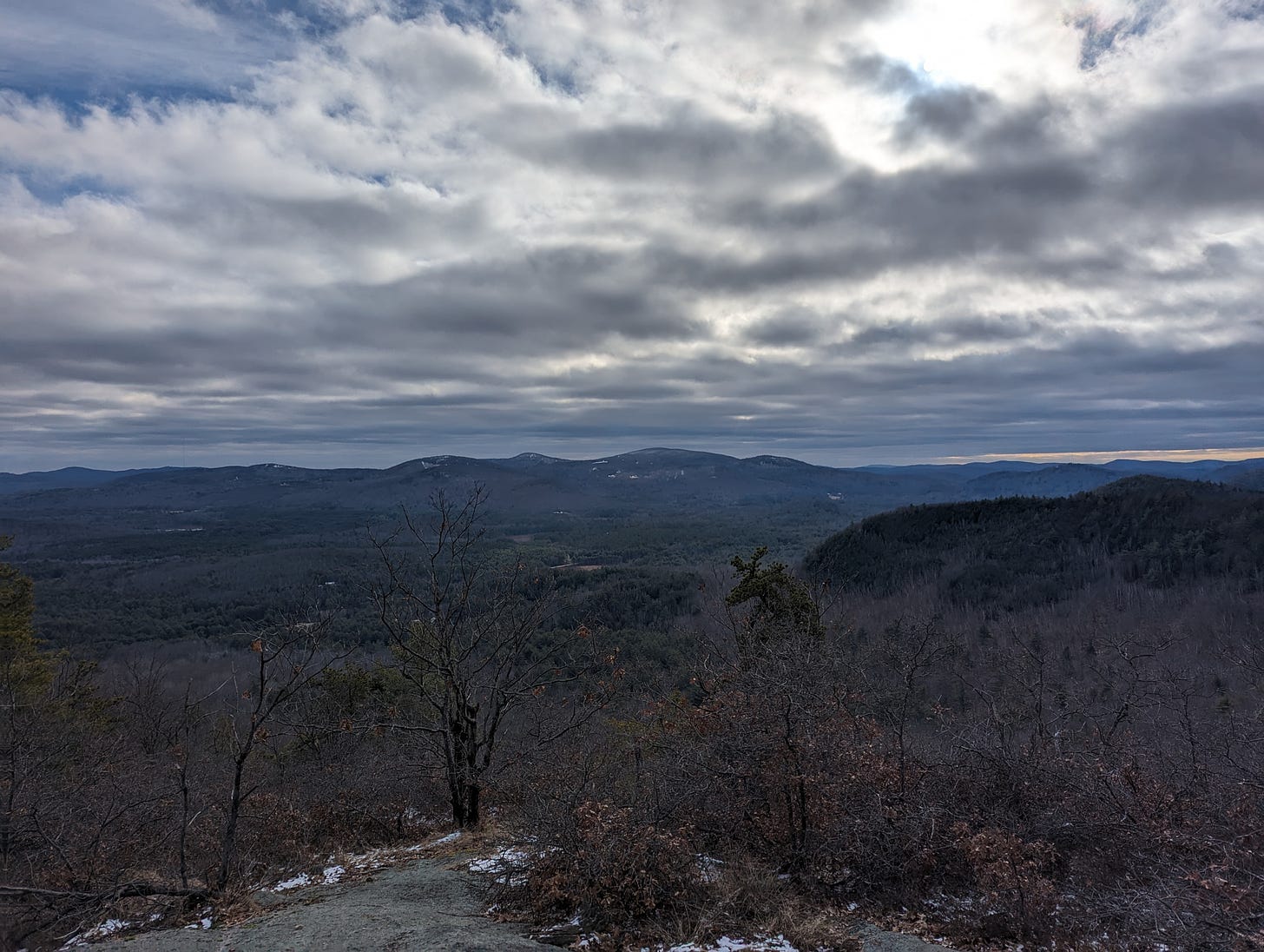 A photo from a scenic overlook, showing heavy clouds, low mountains, and traces of snow on the ground. The sky is heavily overcast.