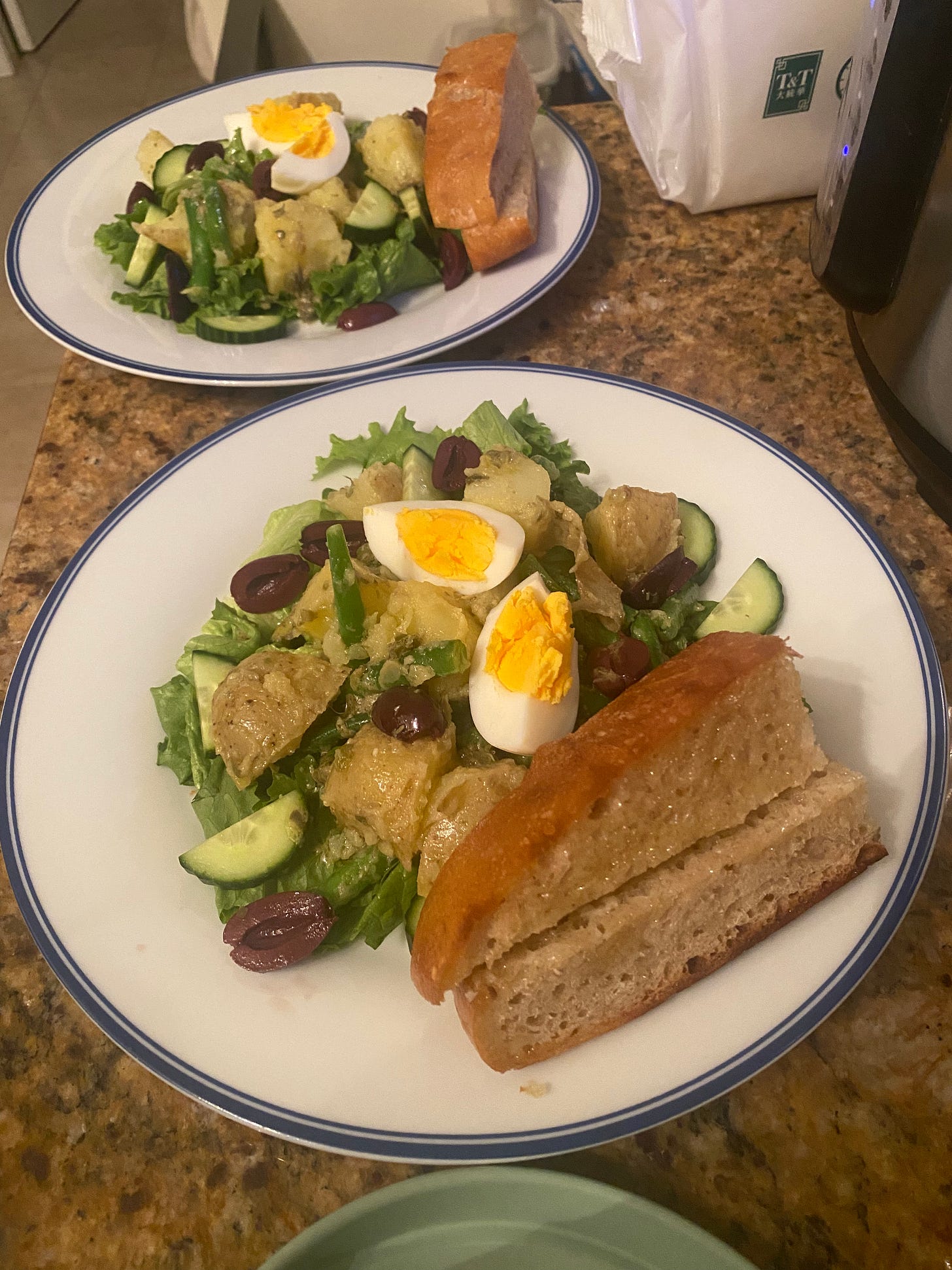 The salad described above, on a white plate with a blue rim. Green lettuces are topped with green beans, potato pieces, cucumber, sliced kalamata olives, and pieces of boiled egg. At the edge of the plate is a slice of garlic bread, cut in half width-wise.