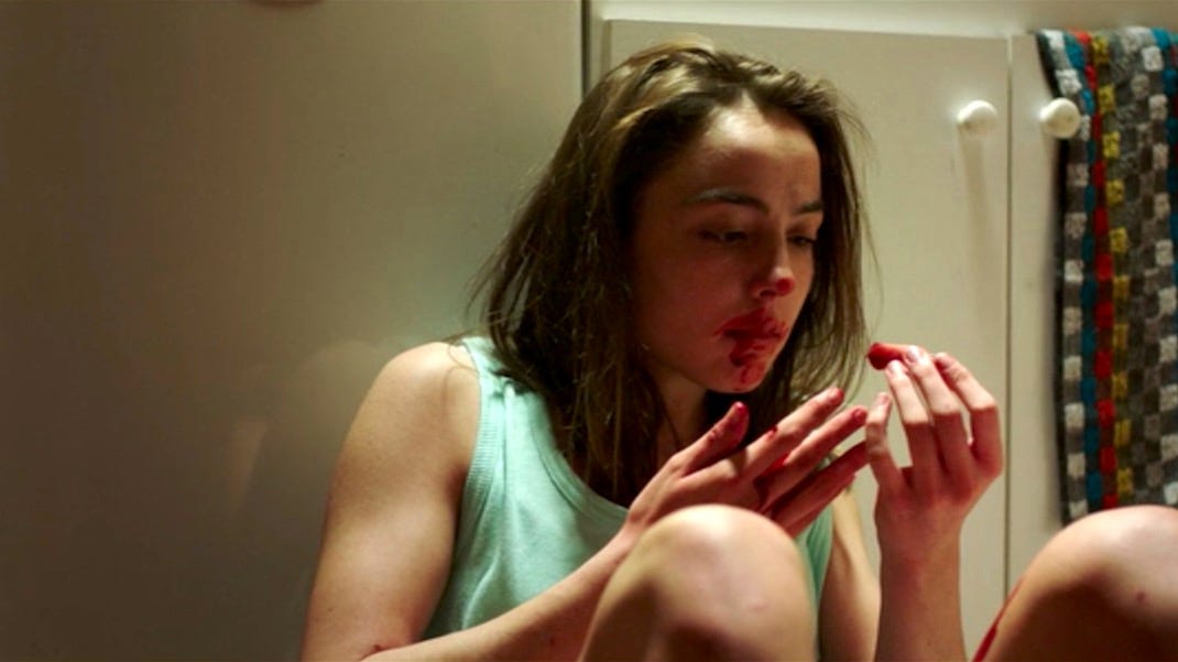 A young white woman with brown hair sitting on the floor with blood on her face holding a bloody finger