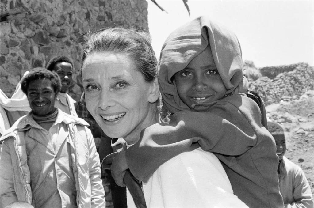 A black and white image of an older Hepburn, standing in a rural African village (I'm afraid I don't know which country) with a smiling child on her back