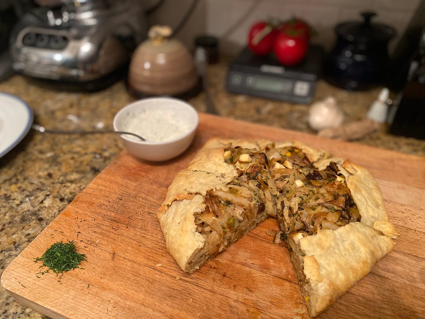 On a wood cutting board, the galette described above, one slice taken out of it at the front, showing caramelized pieces of cabbage dotted with shiitake mushrooms and bits of boiled egg in the mixture. Next to the galette is a little bowl of the sour cream, and a small pile of chopped dill.