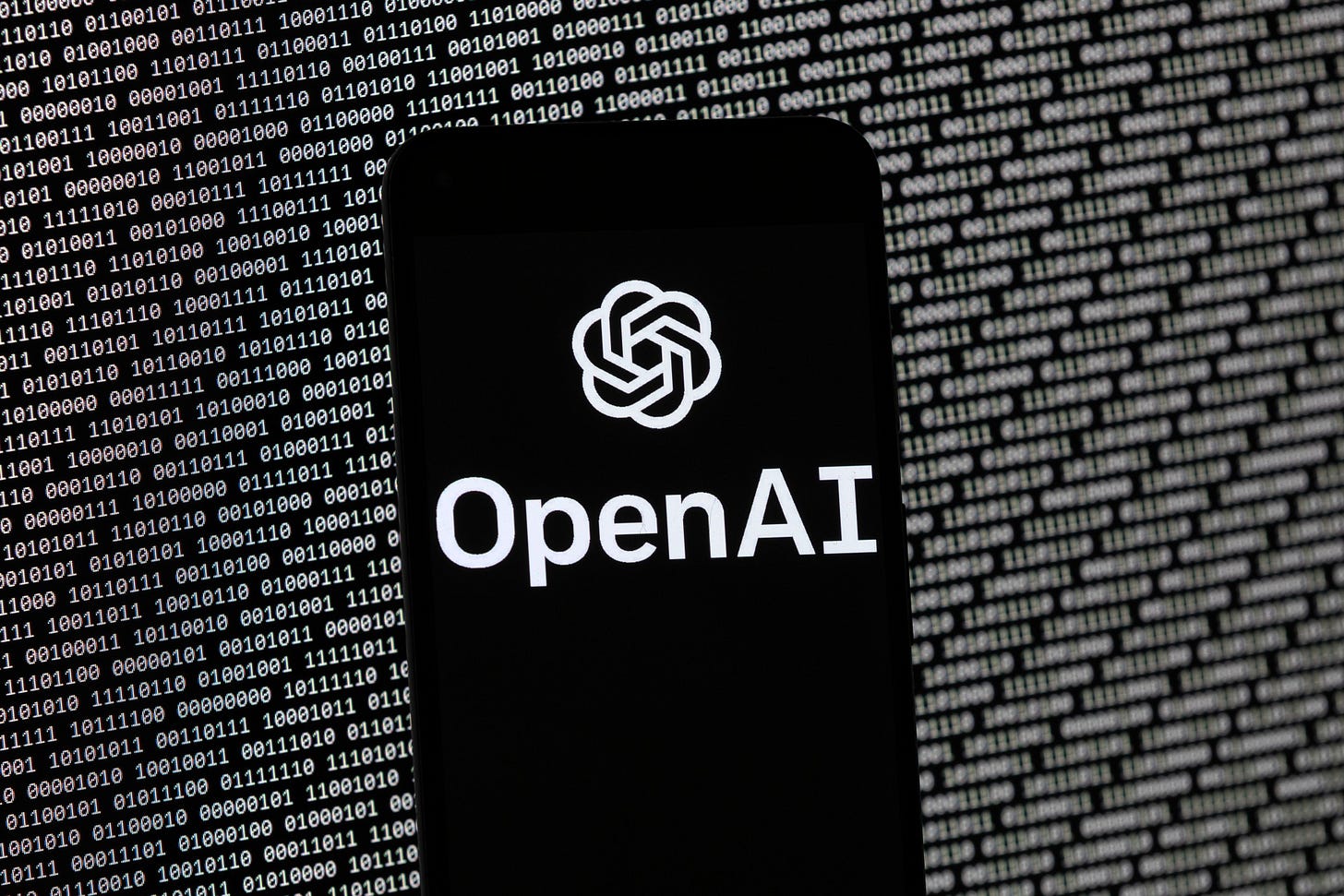 The logo of the Open Ai logo on a mobile device with aa series of zeros and ones in the background on a monitor