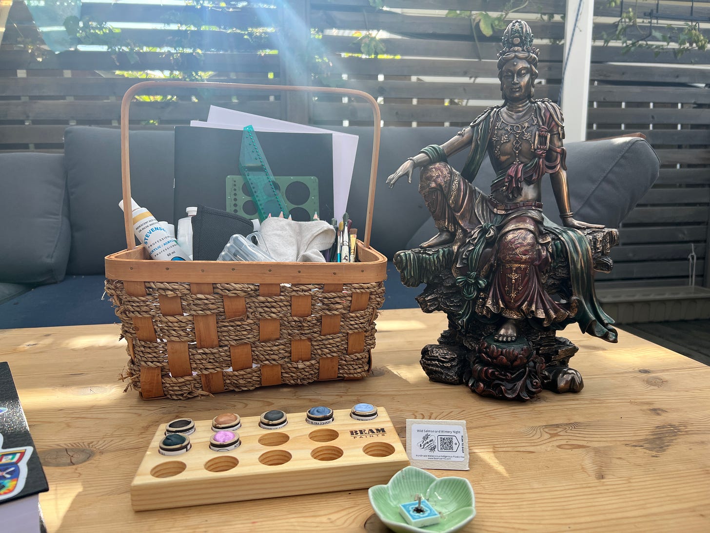 A statue of Kannon, the Japanese depiction of Avalokiteshvara, sitting on a wooden table next to the basket where I keep the supplies I am using for the piece. In the foreground are some watercolours, made by Beam paints, and an incense dish shaped like a flower. 