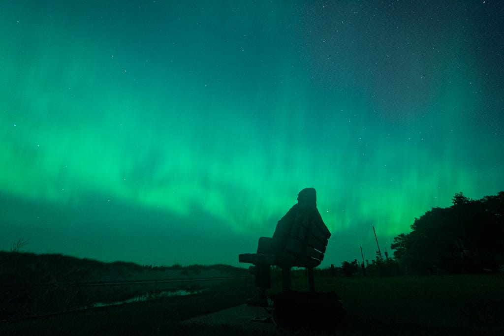 A person sits on a bench, silhouetted by northern lights.