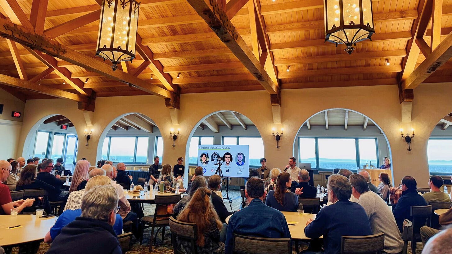 Photograph of the live audience at the Ventech “AI Impacts & Implications” Summit, held in a vaulted, wood-beamed venue with ocean views in the background. A panel of speakers is seated at the front of the room beside a large display screen showing headshots and names of featured panelists: David Wang, Heike Schirmer, and Dr. Paul Leonardi. The discussion is moderated by Mark Weeks. Attendees are seated at round tables, listening attentively to a conversation on the strategic and organizational implications of artificial intelligence. Photograph of the live audience at the Ventech “AI Impacts & Implications” Summit, held in a vaulted, wood-beamed venue with ocean views in the background. A panel of speakers is seated at the front of the room beside a large display screen showing headshots and names of featured panelists: David Wang, Heike Schirmer, and Dr. Paul Leonardi. The discussion is moderated by Mark Weeks. Attendees are seated at round tables, listening attentively to a conversation on the strategic and organizational implications of artificial intelligence.