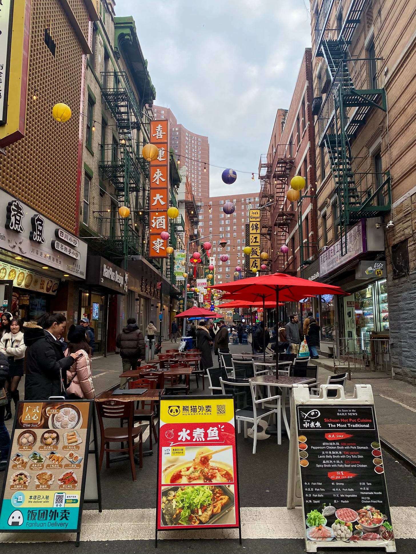 A lively street in Chinatown. There are cafe tables with red umbrellas set up in the road. A string of lights with paper lanterns is suspended over the street, crisscrossing between the two buildings. People are sitting enjoying food and tea, milling between storefronts, and checking phones. The blue of the sky glows from behind thick clouds.