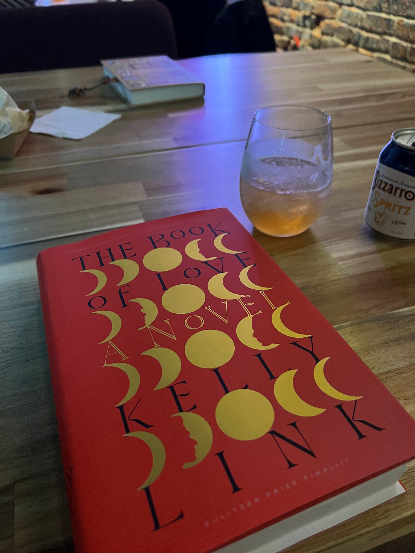 A copy of The Book of Love sitting on a wooden table in a bar next to a white wine spritzer in a glass. 