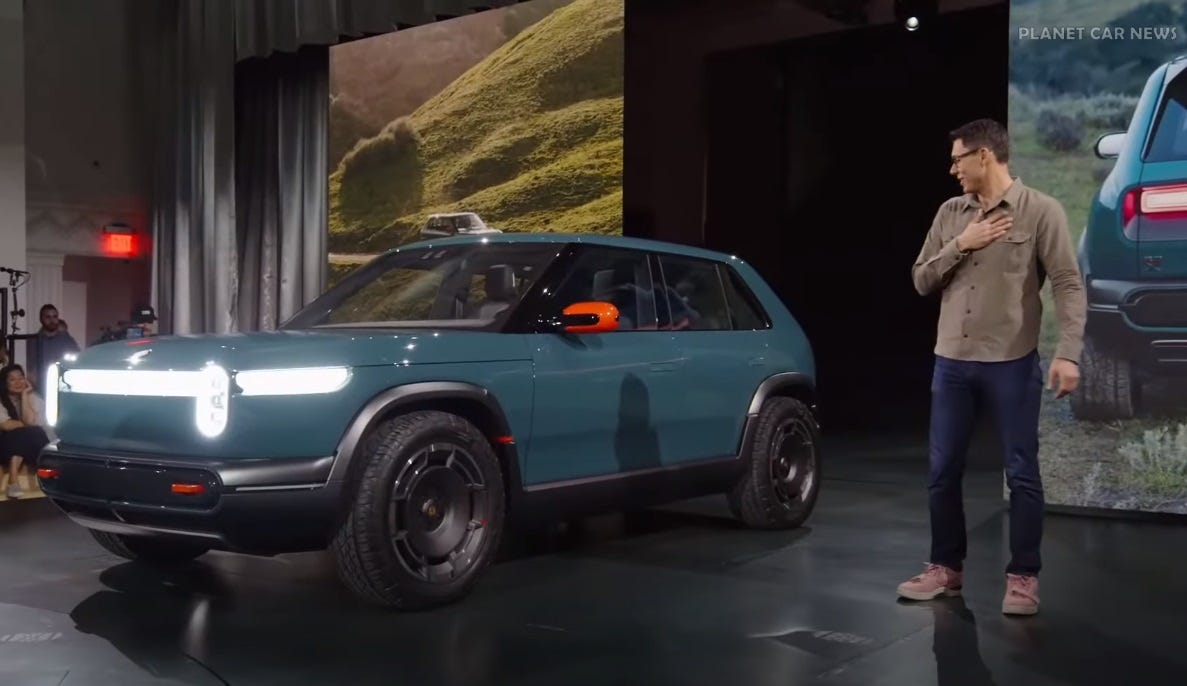 Rivian CEO RJ Scaringe, in jeans and an untucked tan shirt, admires a green prototype EV on stage 