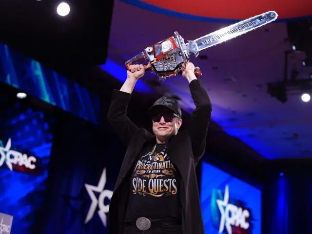 Elon Musk holding a chainsaw above his head with both hands. He is wearing a black hat, a black jacket, and a black t-shirt.